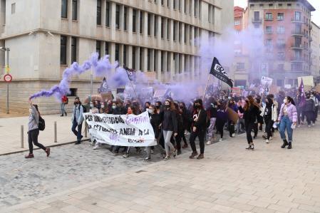 Pla general de la capçalera de la manifestació feminista convocada pel SEPC passant pel carrer de Pompeu Fabra de Girona tirant fum de color lila el 8 de març de 2021 (Horitzontal)